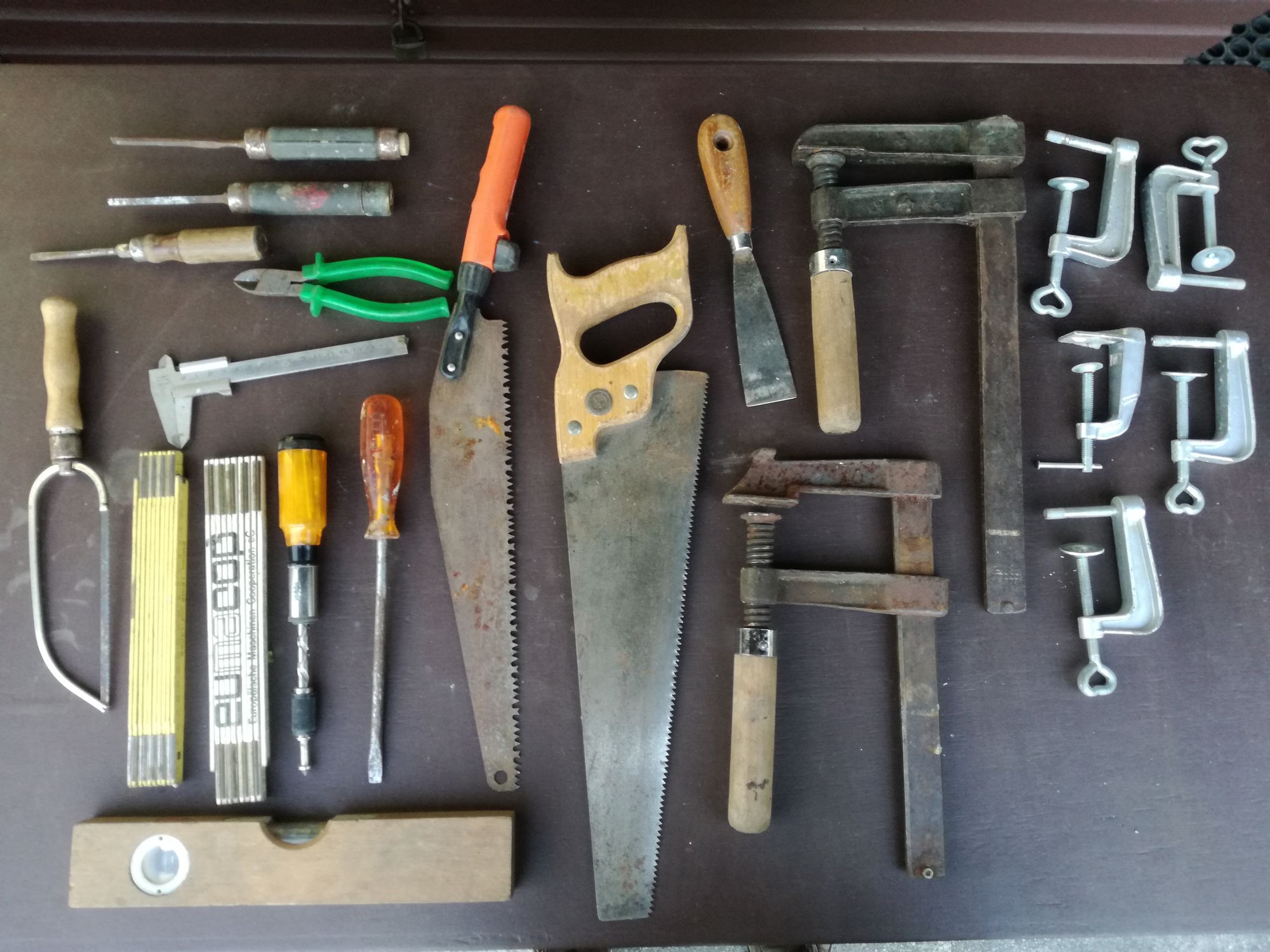 A variety of well-used tools laid on a solid table. Screwdrivers to the top left, pliers, calipers, several kinds of saw, a level, two large clamps, five smaller clamps, and a scraper.